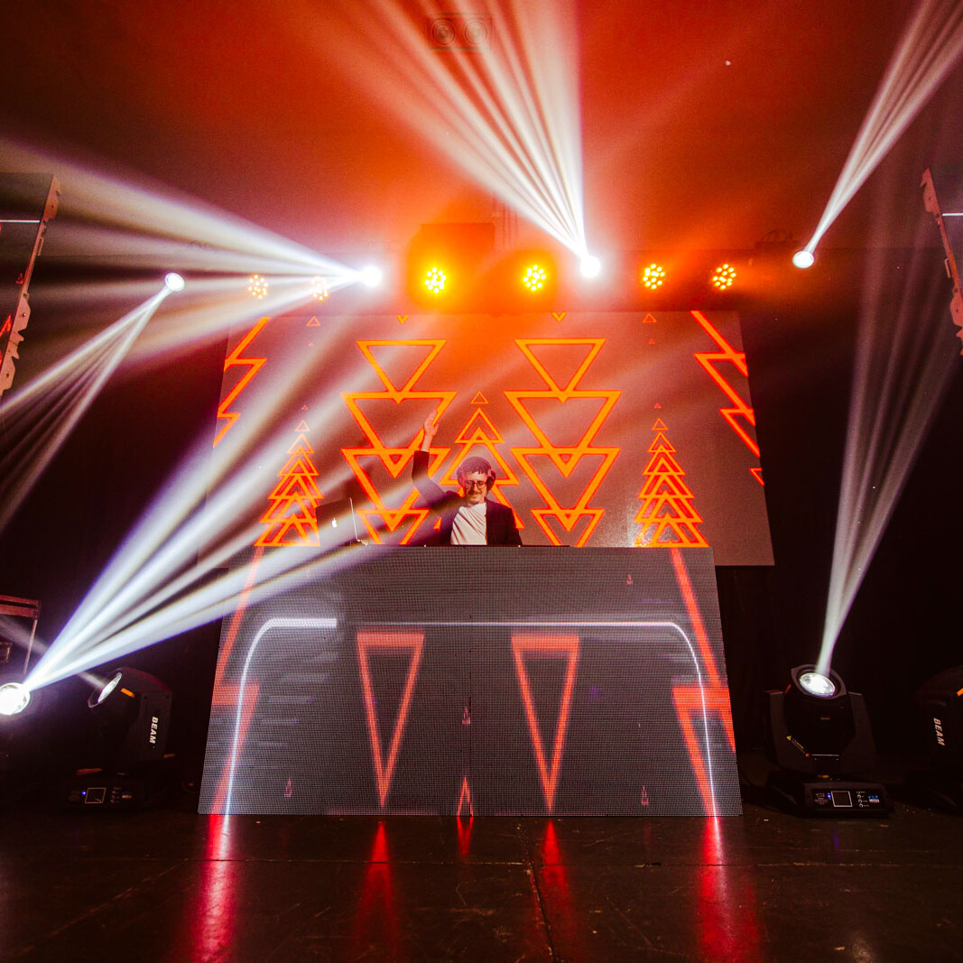 DJ auf Bühne mit roter LED-Videowand, geometrischen Mustern und starken Lichtstrahlen bei einem Festival oder Club-Event
