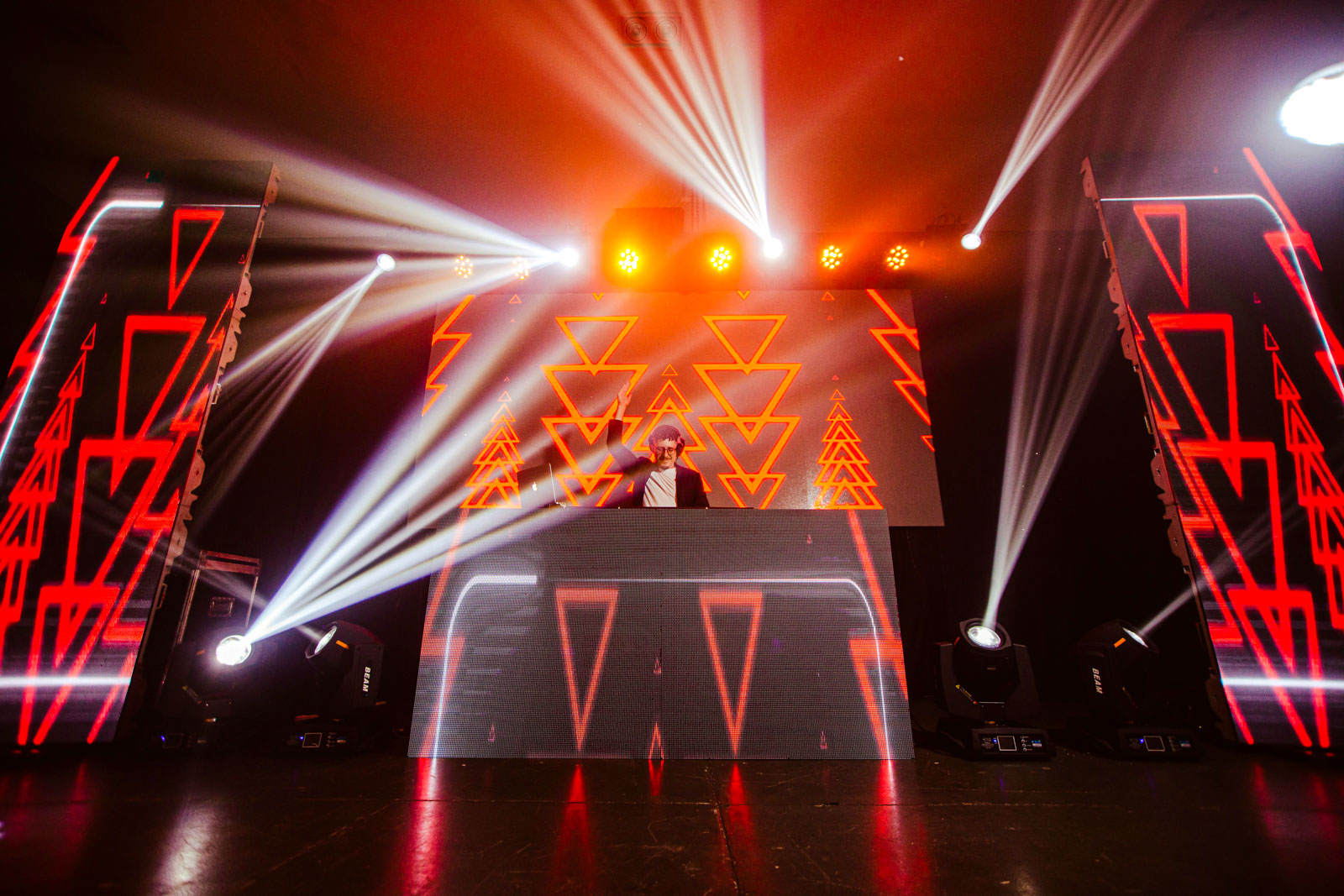 DJ auf Bühne mit roter LED-Videowand, geometrischen Mustern und starken Lichtstrahlen bei einem Festival oder Club-Event