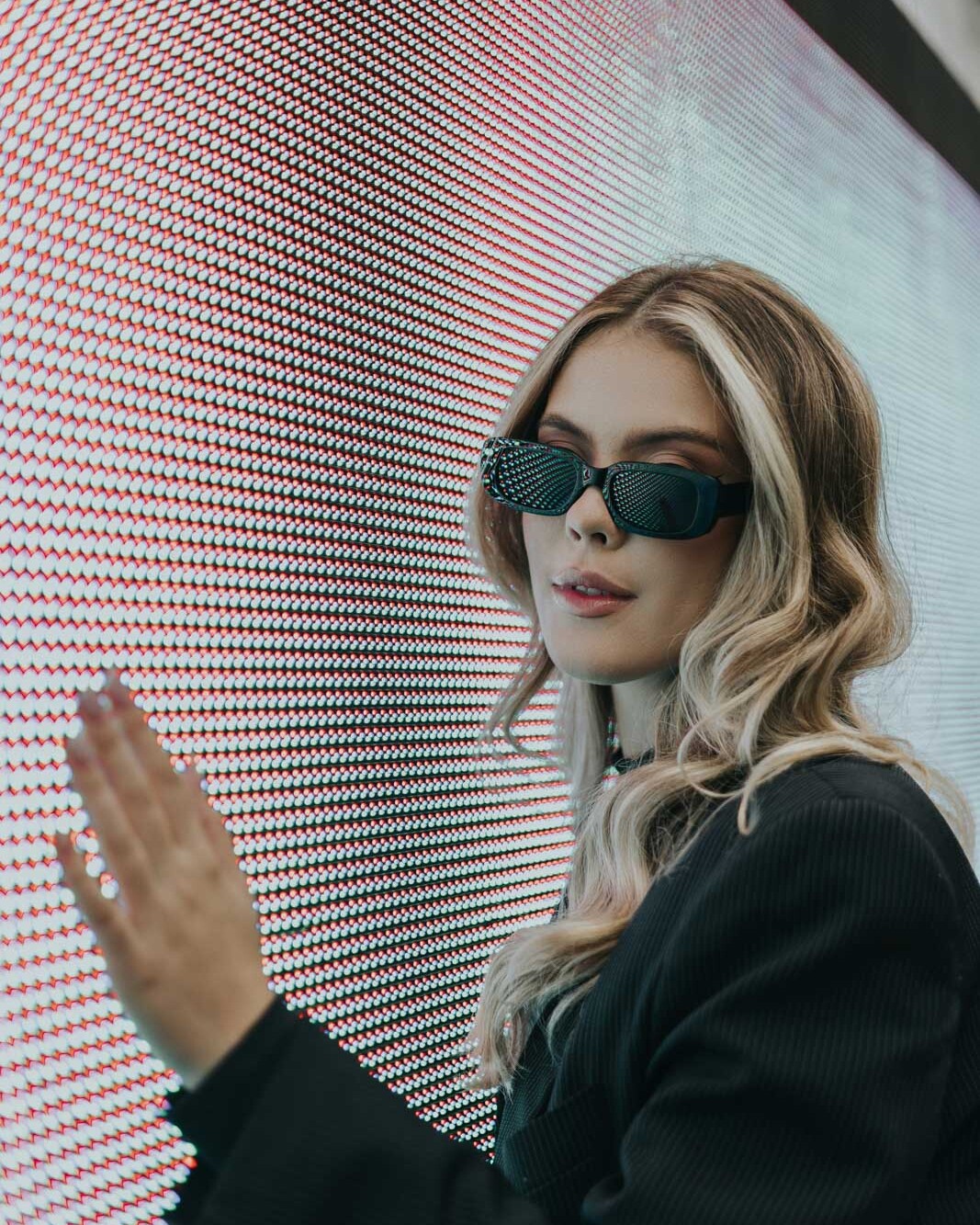 Frau mit Sonnenbrille berührt interaktive LED-Wand mit leuchtendem Pixelmuster bei einem Event oder einer Messe